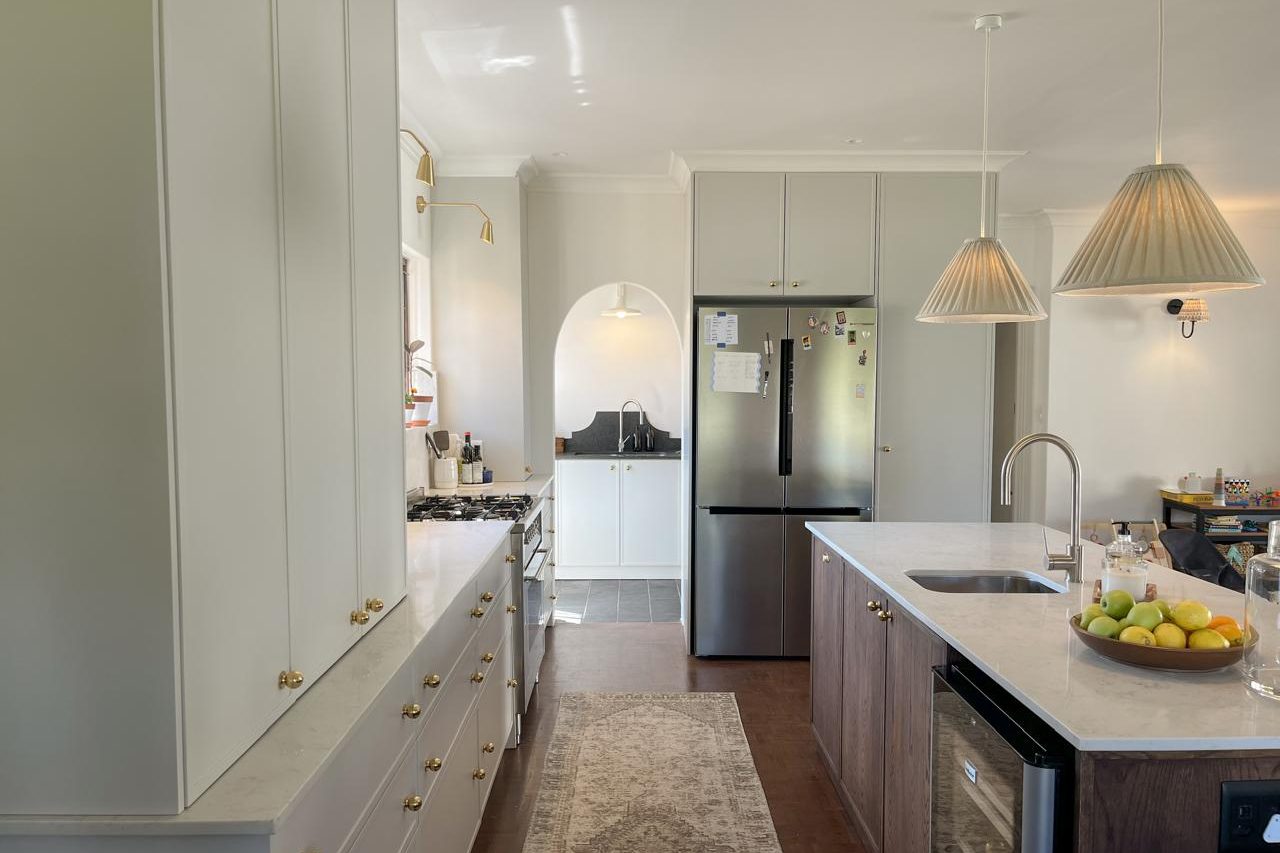 A kitchen with pale painted cabinetry and brass knobs, an arched doorway to a scullery, and a timber island with a stone top